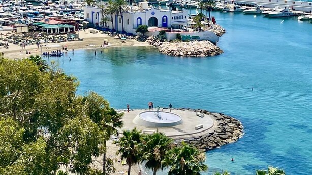 Un port de plaisance avec des bateaux amarrés, une plage de sable et une zone circulaire centrale agrémentée d’une fontaine.
