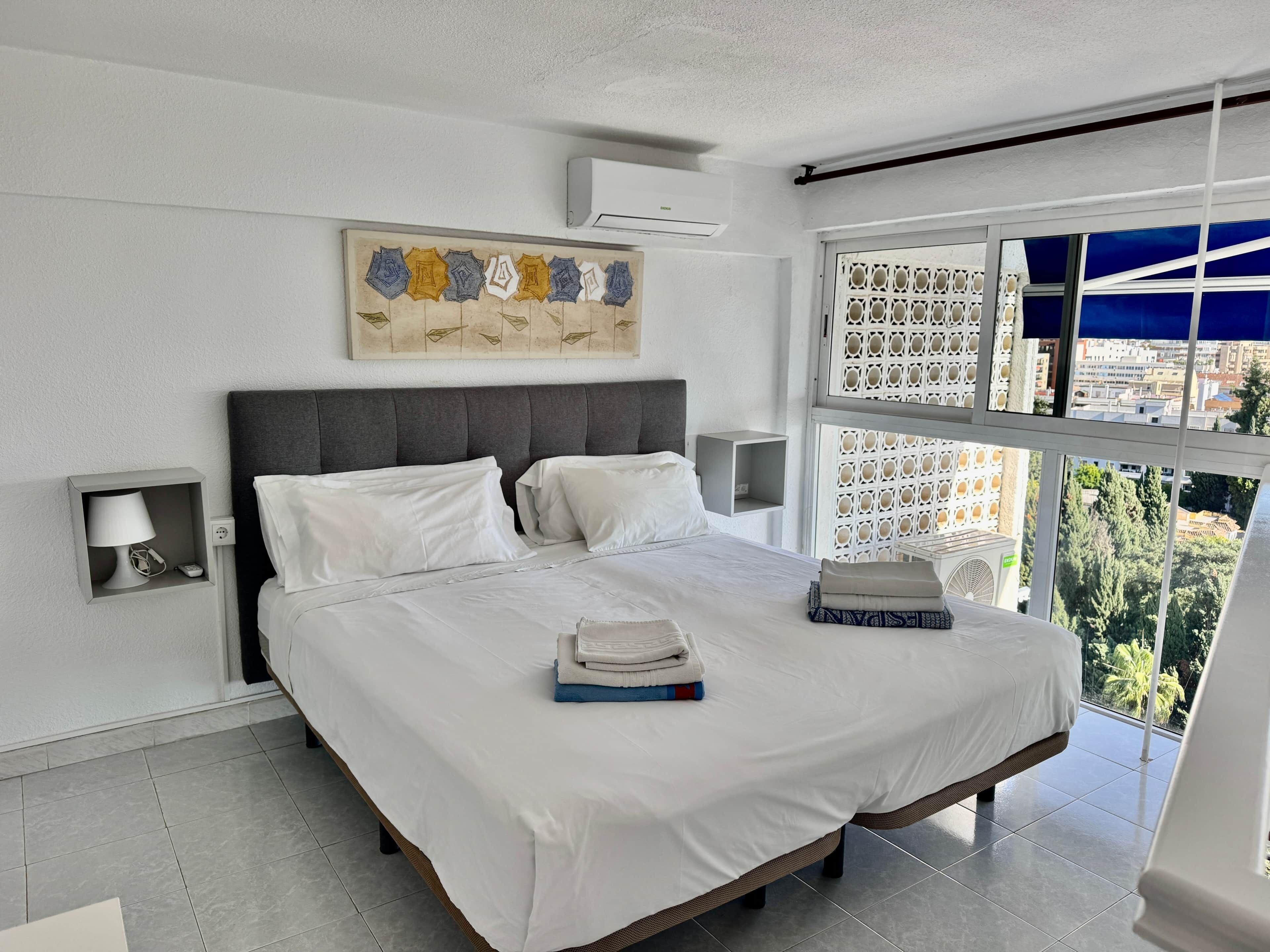 Une chambre moderne avec un grand lit, une table de chevet, un climatiseur fixé au mur et une fenêtre donnant sur la ville.
