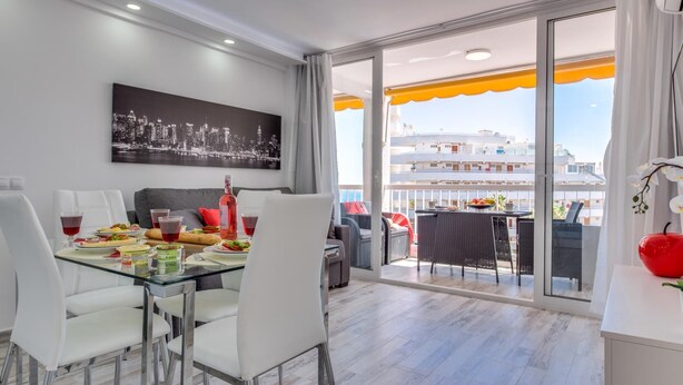 A modern dining area with a glass table, white chairs, and a cityscape view through large windows.