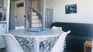 A room with a round table, white chairs, a dark sofa, a staircase, and a painting on the wall. - Duplex 5 people Carnac Beach with Feet (Carnac)