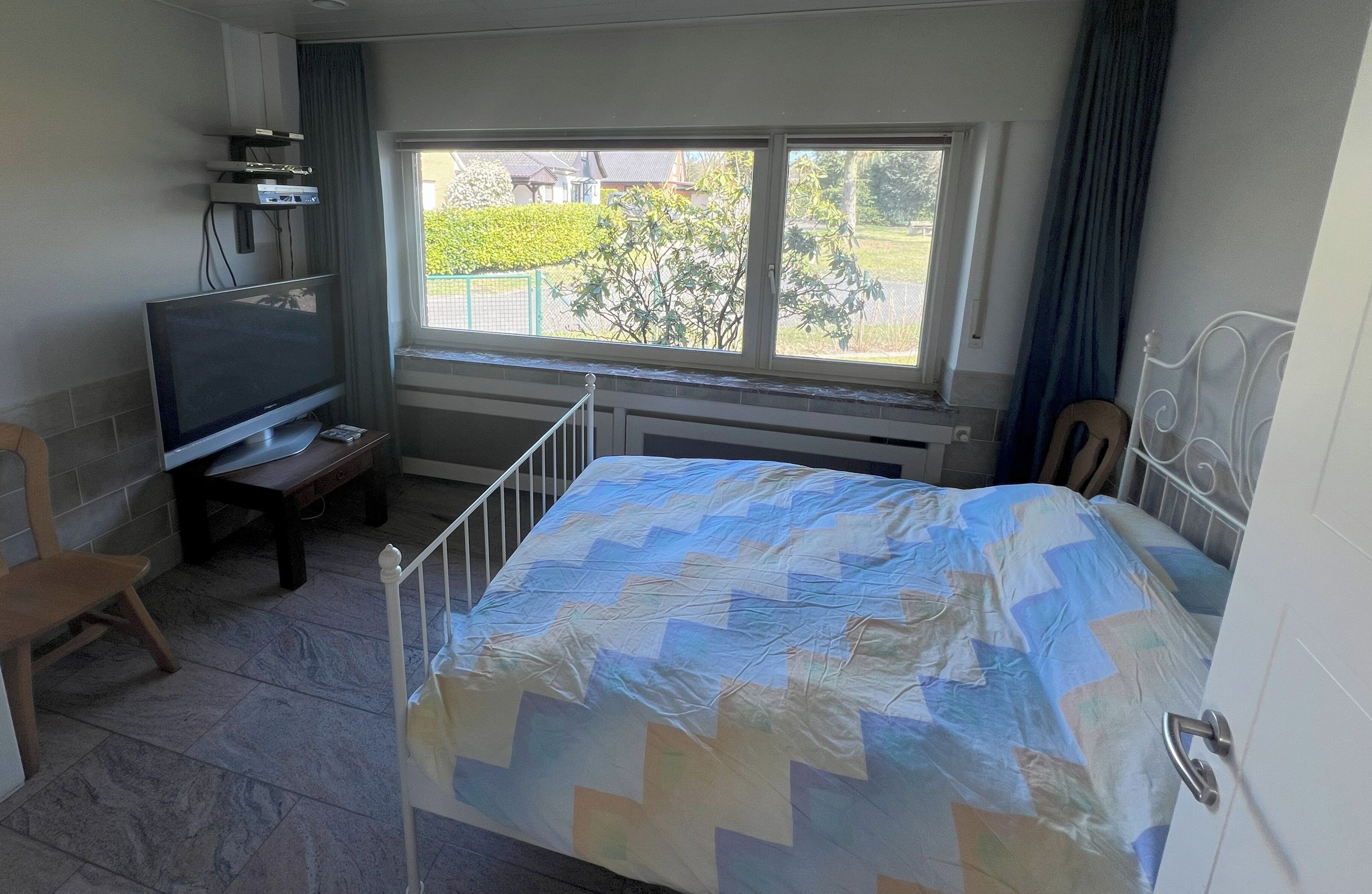 Quarto com cama, televisão, cadeira e janela com vista para área verde.