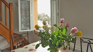 A dining table with a floral tablecloth, a potted plant with roses, and a view of a building through an open window. - Comfortable duplex on the peninsula of Giens facing Porquerolles sea view (La Tour Fondue)