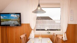 Zona de menjador amb taula preparada per a dues persones, televisió penjada a la paret i finestra amb cortines.