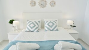 Una cama bien hecha con dos almohadas, una colcha azul y dos toallas enrolladas. Hay dos mesitas de noche con lámparas y pequeñas macetas. La habitación tiene una decoración en tonos blancos y espejos decorativos en la pared.