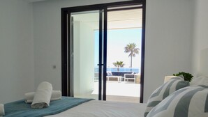 Una habitación de hotel con una cama, una toalla azul, un almohadón a rayas y vistas a un balcón con mobiliario exterior y una palmera.