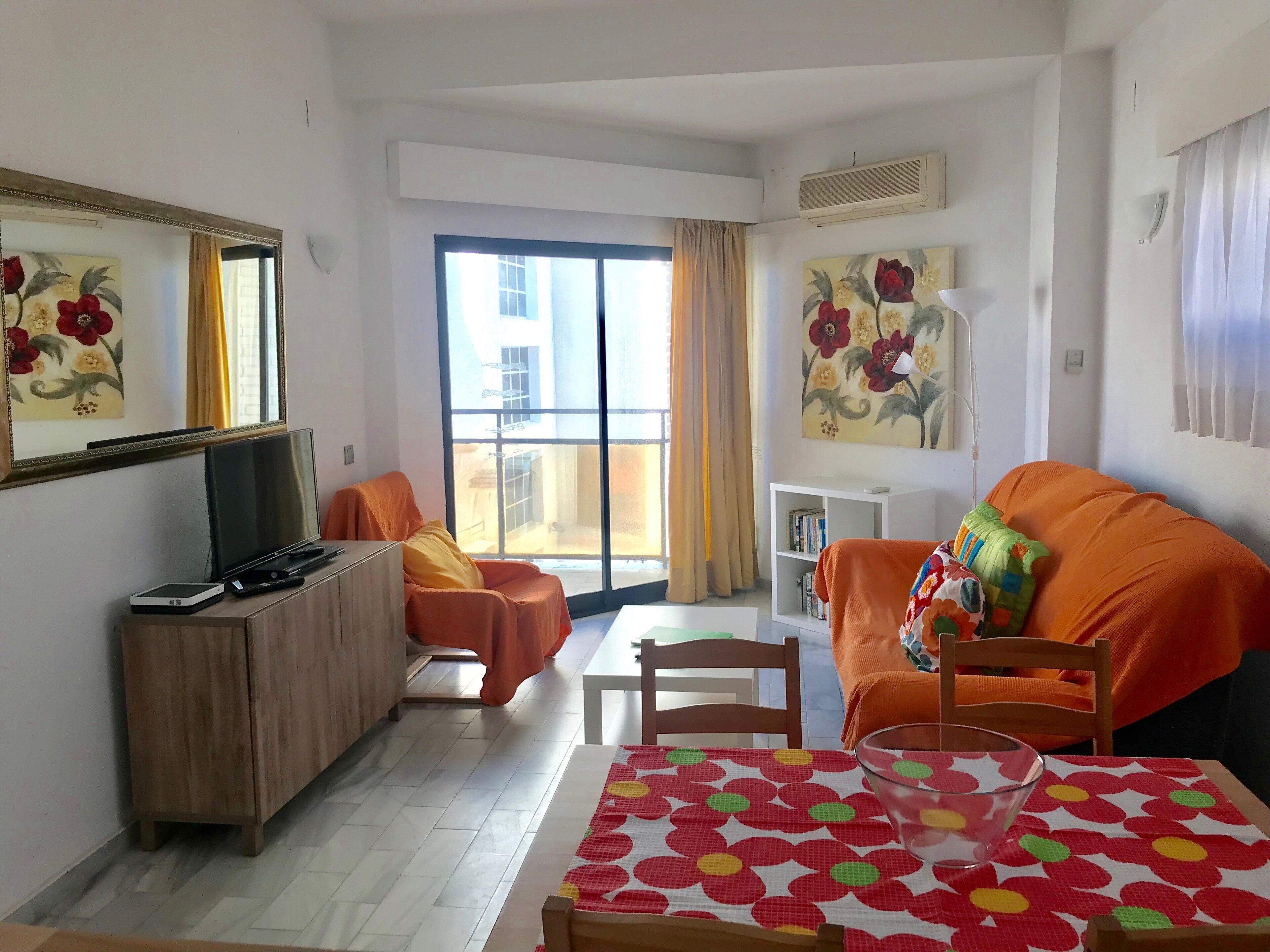 A hotel room with a large window, a sofa, a dining table with a patterned tablecloth, a TV on a wooden cabinet, and artwork on the walls.