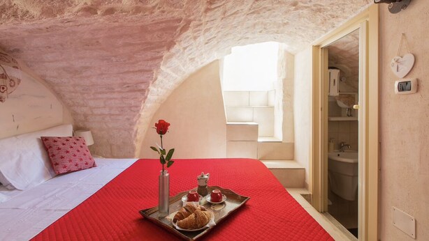 Une pièce chaleureuse avec une couverture de lit rouge, un plateau contenant des croissants et une rose, et une salle de bain visible à travers une porte ouverte.