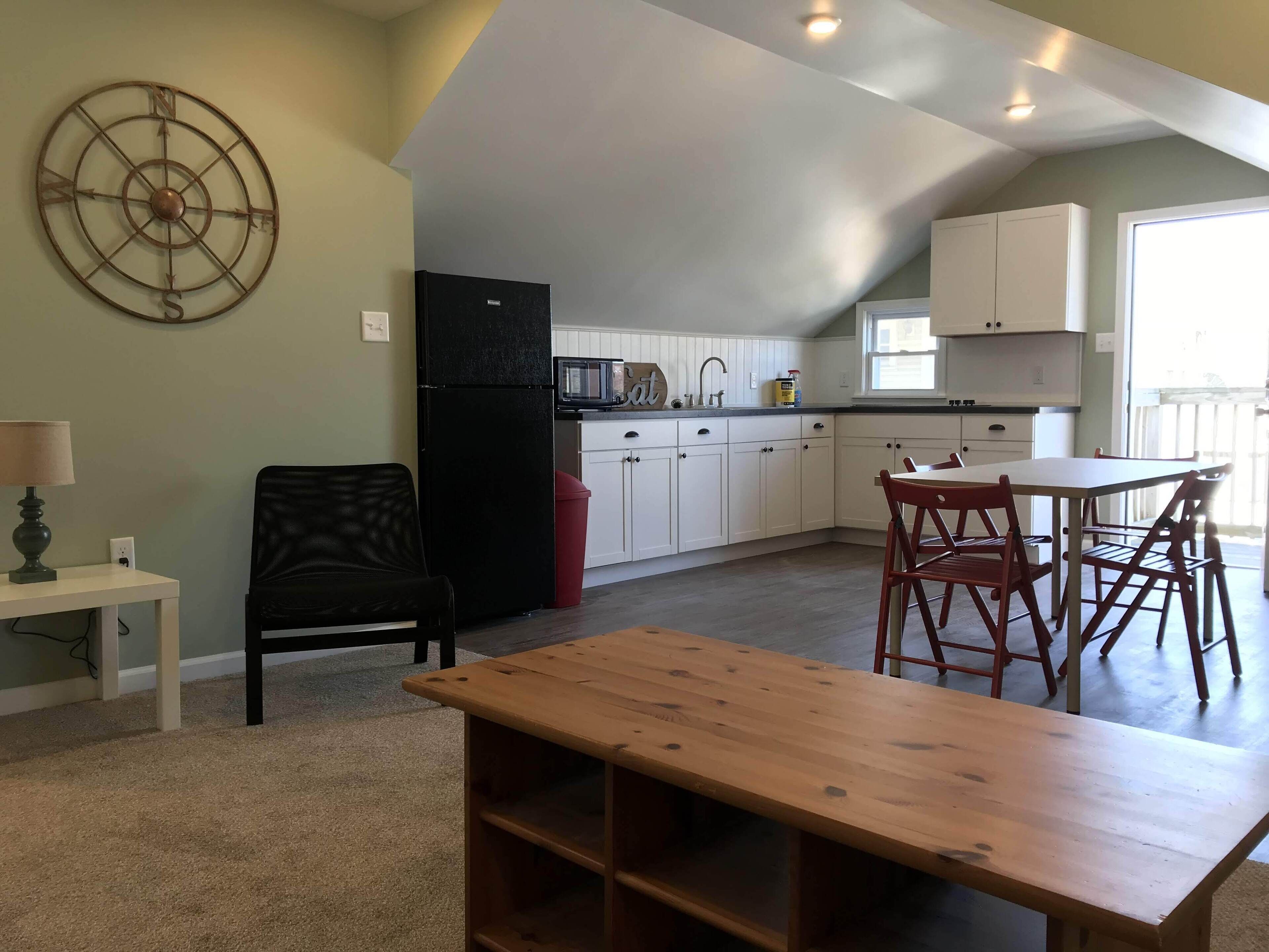 A compact kitchen with white cabinets, a black refrigerator, and a microwave. There is a dining area with a wooden table and red chairs.