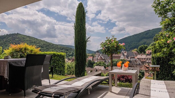 A terrace with a lounge chair and table, overlooking a village with traditional houses and a mountain backdrop.