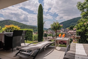 Eine Terrasse mit Liegestuhl und Tisch, mit Blick auf ein Dorf mit traditionellen Häusern und einem Berg im Hintergrund.