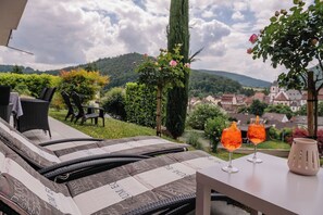 A lounge area with a view of a village and hills, a table with two glasses of orange drink, and a wicker sofa with a patterned cushion. - Beautiful apartment Casa Rossa in Neckarsteinach am Neckar. (Neckarsteinach)