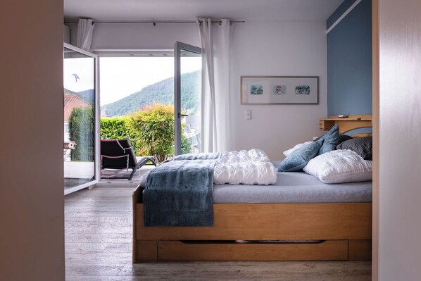 Ein Schlafzimmer mit einem hölzernen Bett, Blick auf Grünflächen und Hügel, sowie ein Sessel am Fenster.