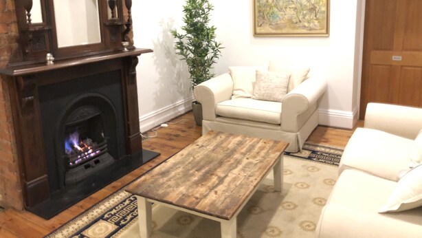 A living room with a fireplace, a white sofa, a wooden coffee table, and a painting on the wall. - Napier Cottage - Cozy Hills Retreat (Hawthorndene)