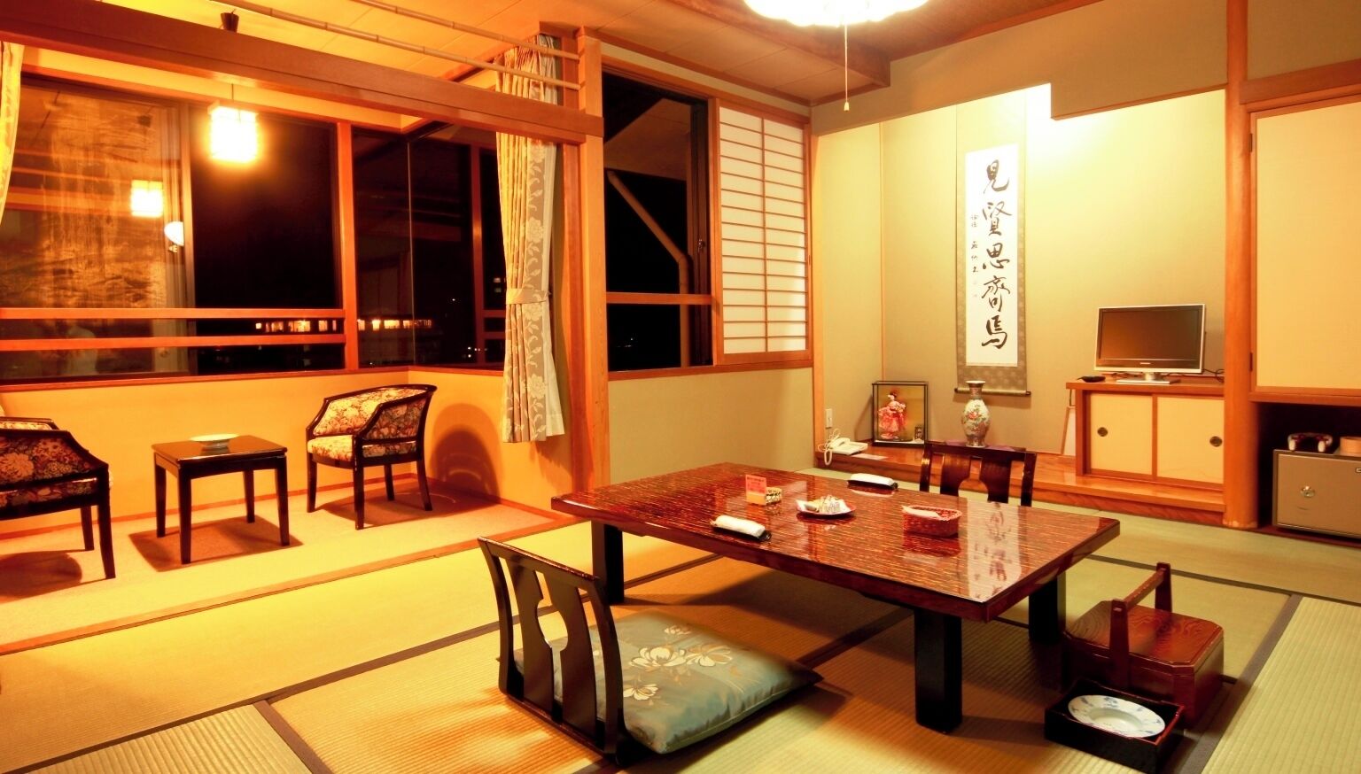 Ein traditioneller japanischer Raum mit Tatami-Boden, niedrigem Tisch, Stühlen, einem Fernseher und einer Schiebetür.