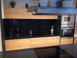 Een moderne keuken met een zwart aanrecht, houten kastjes, een glazen spatwand, een inbouwoven en een koffiezetapparaat.