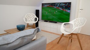 A living room with a television displaying a soccer game, two white chairs, a blue bowl, and a grey sofa. - Beautiful new, modern apartment in Grömitz, 43sqm (Grömitz)