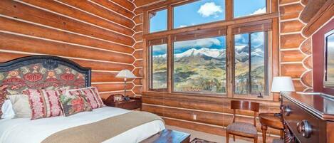 A log cabin room with a bed, a chair, a dresser, and a large window with a mountain view.