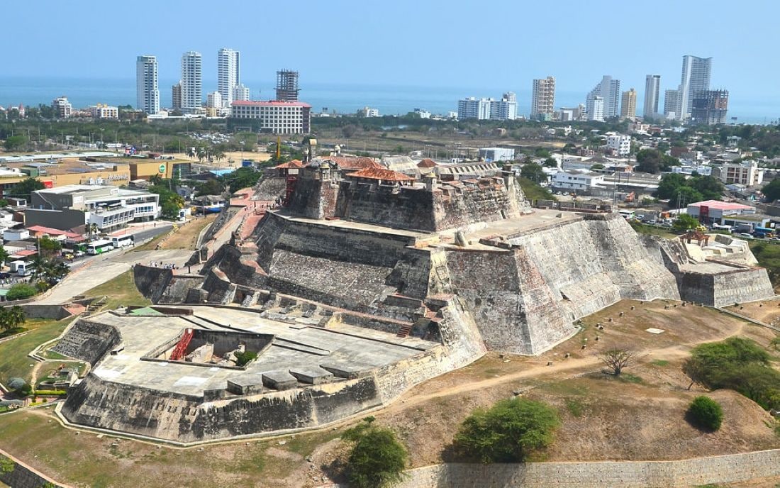 Vista aerea di un'antica fortezza con numerose mura e un fossato, circondata da edifici moderni e un cielo sereno.