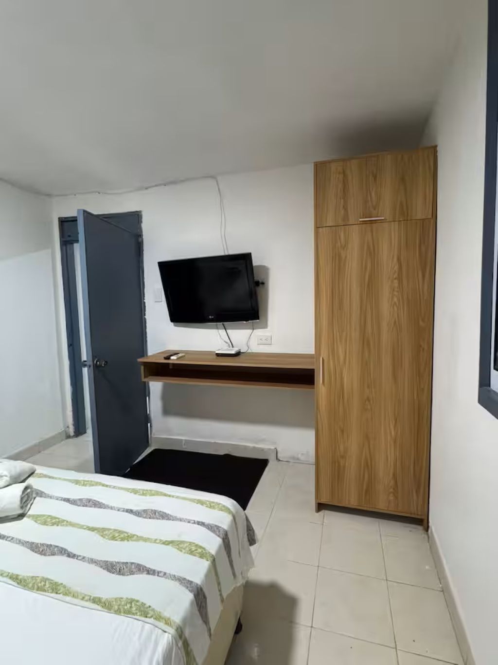 A bedroom with a bed, a TV mounted on a wall, a wooden wardrobe, and a door.