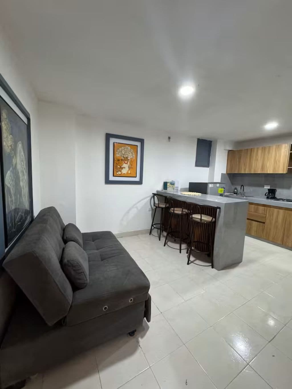 Una sala de estar moderna con un sofá gris, una zona de cocina con armarios de madera y un juego de taburetes de bar.
