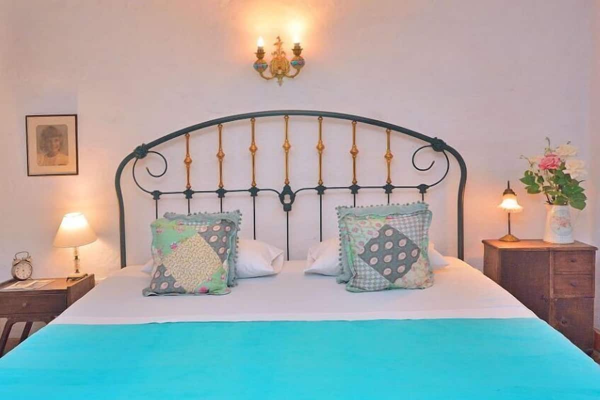 A neatly made bed with a teal bedspread, two decorative pillows, a wall-mounted light fixture, and a small bedside table with a lamp.