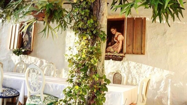 A rustic outdoor dining area with a table and chairs, surrounded by greenery and a stone wall.