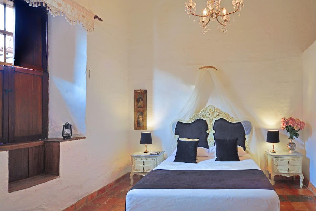 A bedroom with a large bed, a canopy, two bedside tables with lamps, a nightstand with a vase of flowers, and a window with curtains.