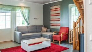 Un salon moderne avec un canapé d’angle gris, un fauteuil rouge, une table basse blanche et un tapis rouge. Un escalier en bois se trouve à droite, et il y a une fenêtre avec des rideaux.