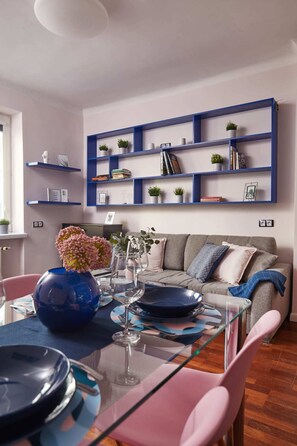 A modern living room with a glass dining table, pink chairs, a grey sofa, and blue shelves with books and plants. - Plac Zbawiciela Hipster Apartment (Warsaw)