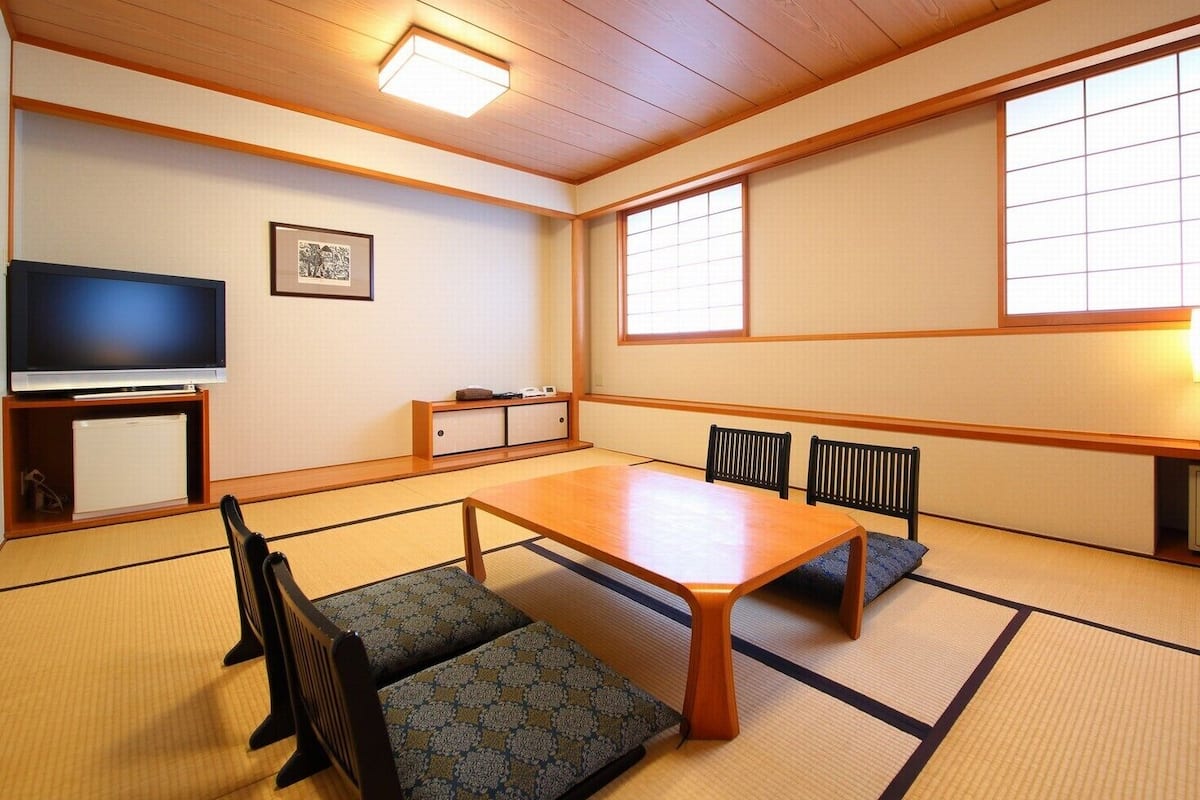 Een traditionele Japanse kamer met tatami vloerbedekking, een lage tafel, stoelen, een televisie en een raam met shoji schermen.