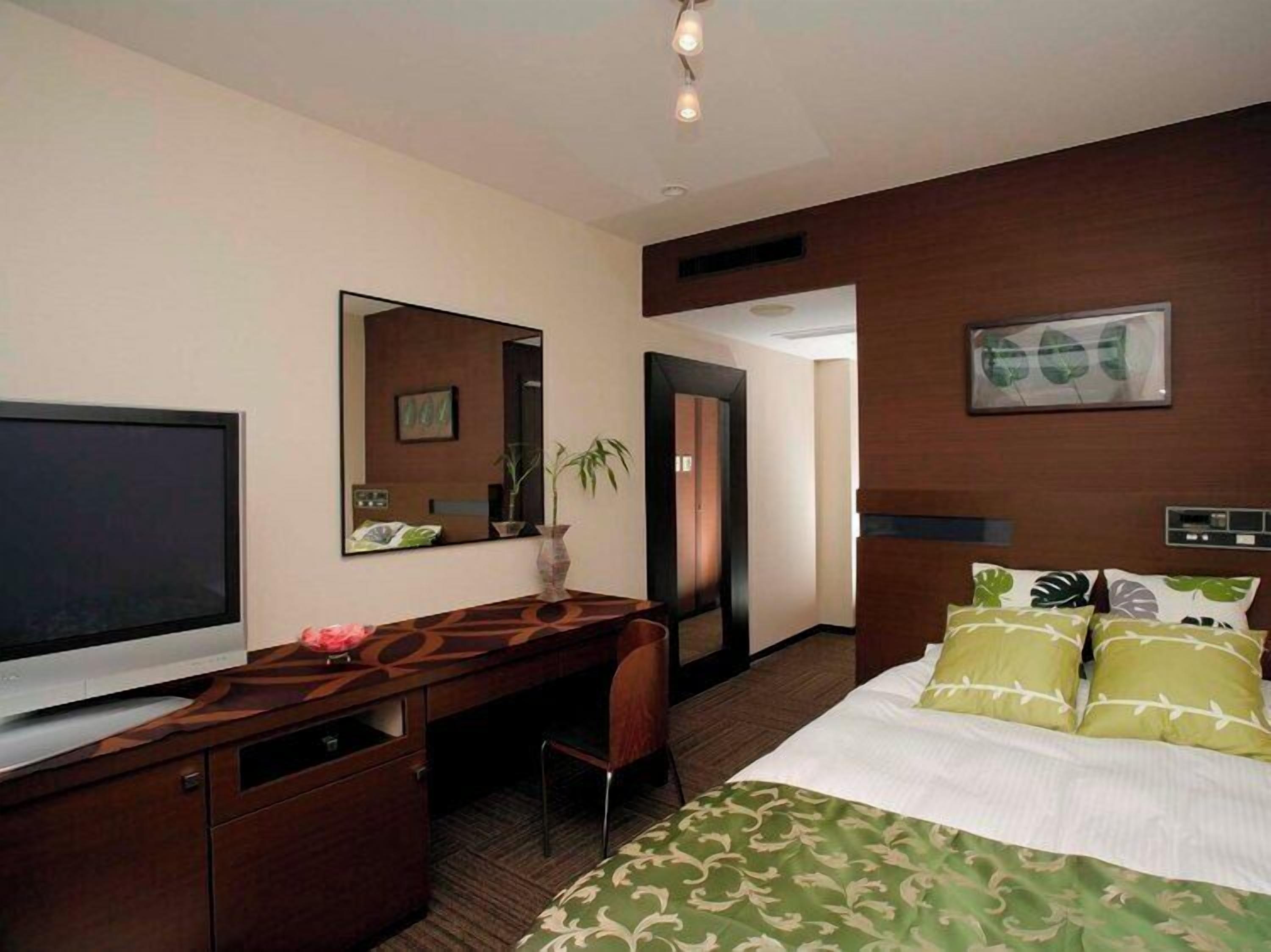A hotel room with a bed, a desk, a TV, a mirror, and a door.
