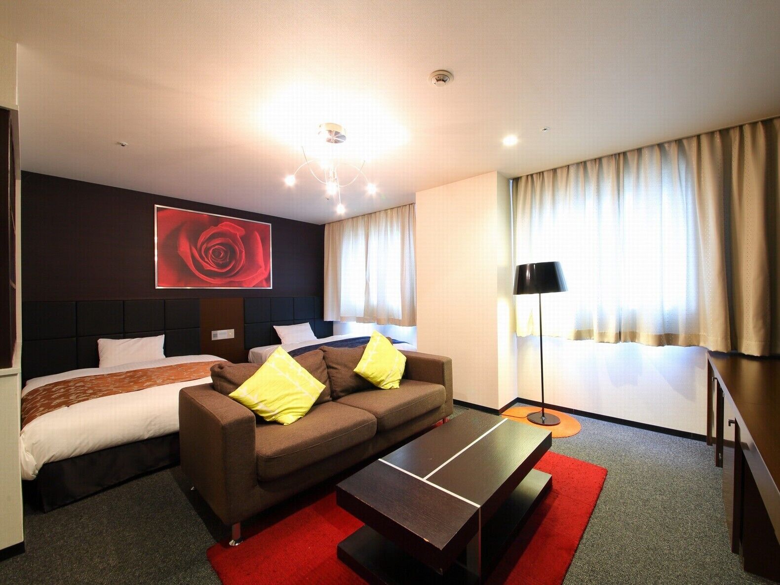 A hotel room with a bed, a sofa with pillows, a coffee table, a red rug, a floor lamp, and a large red artwork on the wall.