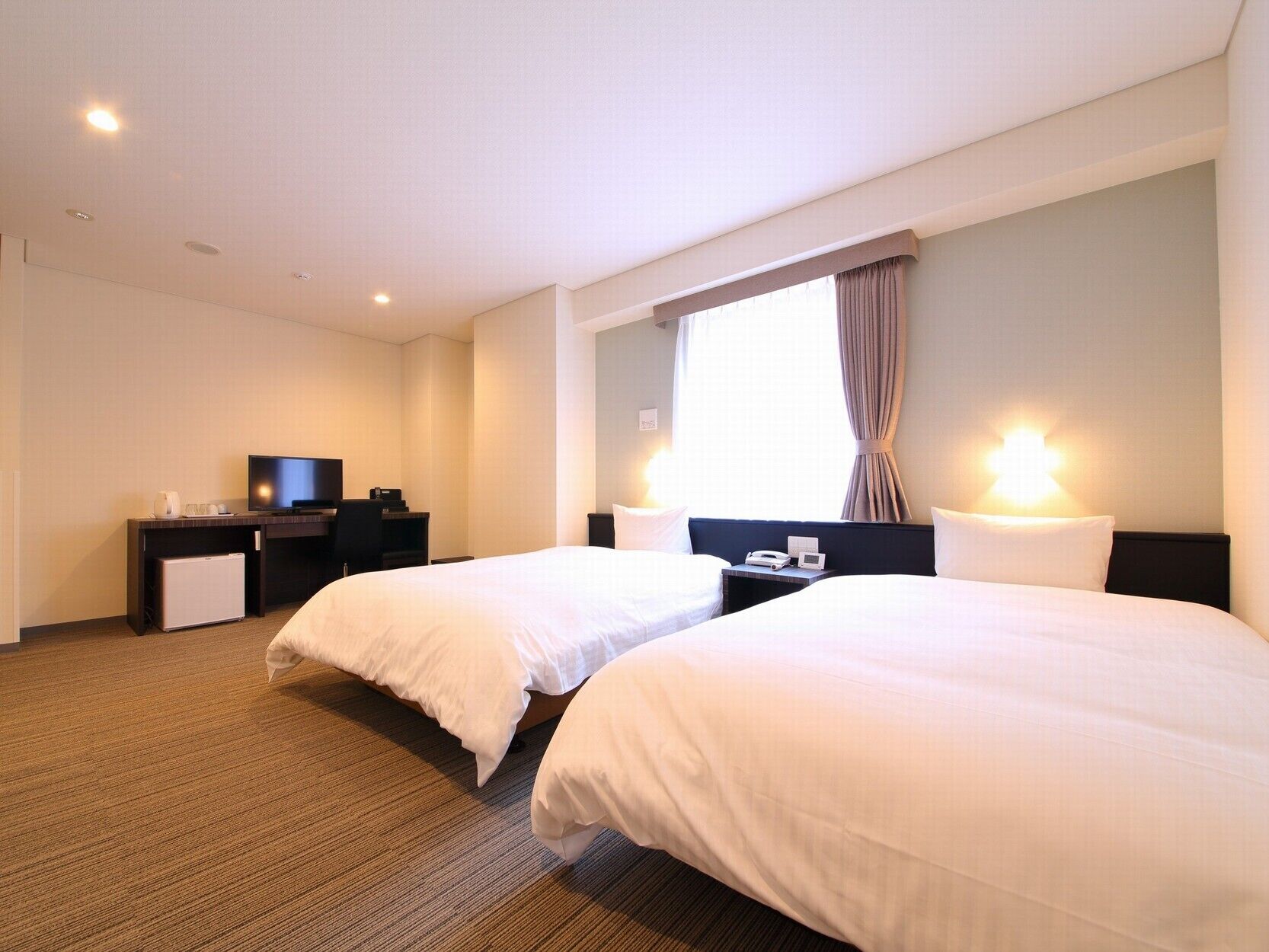 A hotel room with two beds, a TV, a desk with a computer, and a window with curtains.