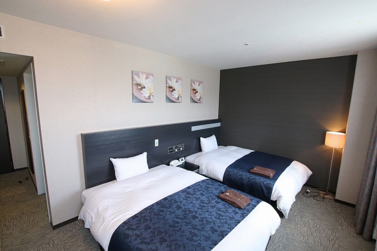Een hotelkamer met twee bedden, een donker hoofdbord, een nachtkastje met een telefoon en een lamp.