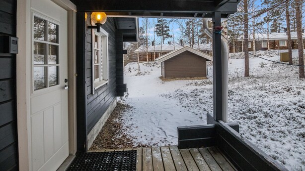 Une cabane en bois avec un porche couvert, une cour enneigée et un petit abri de jardin à l’arrière-plan.