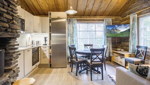 Ein gemütliches Blockhaus-Interieur mit Holzbalkendecke, Steinwand, moderner Küche, Essbereich und einem an der Wand befestigten Fernseher.