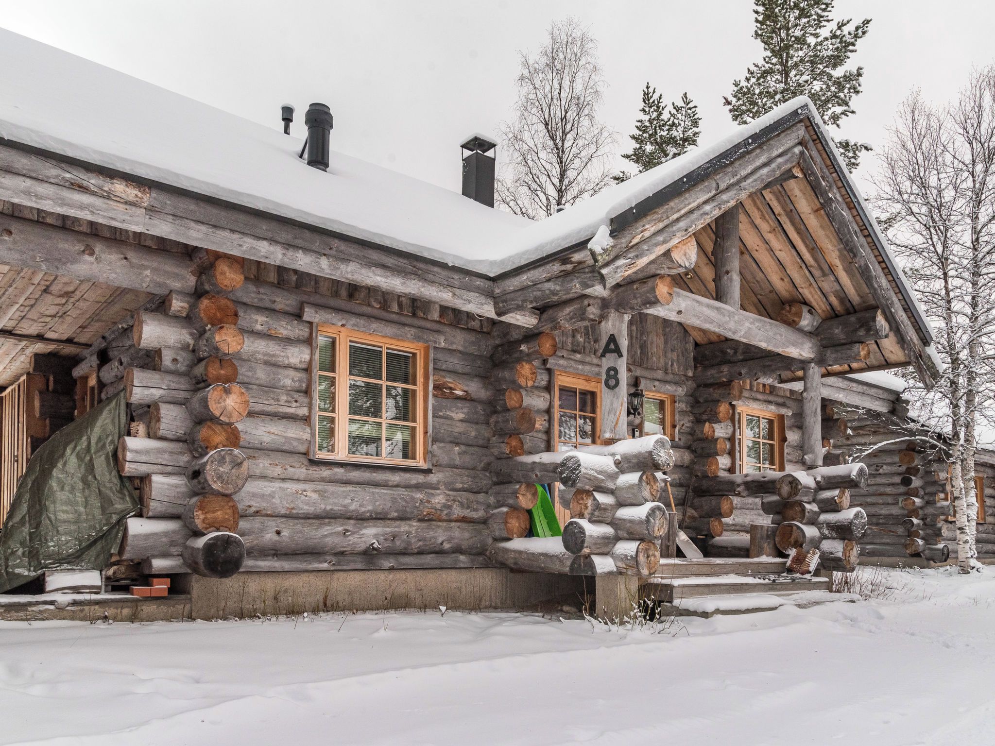 Une cabane en rondins avec un toit enneigé, des bûches empilées à l’extérieur et une cheminée.