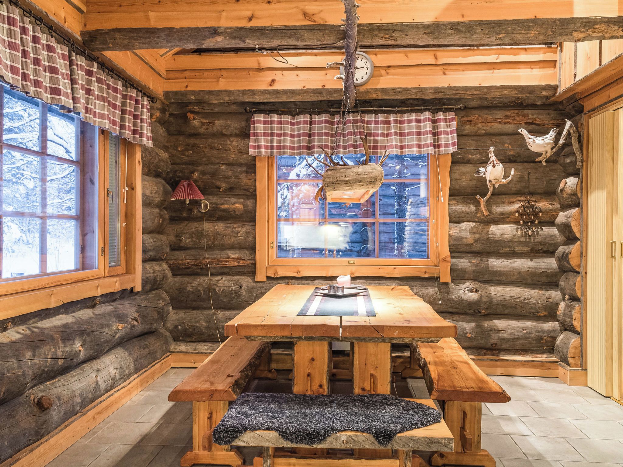 A rustic log cabin interior with a wooden table, plaid curtains, and a window with a view of snowy mountains.