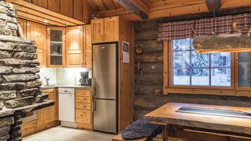 A log cabin interior with a stone wall, wooden cabinets, a stainless steel refrigerator, and a window with a view of snow-covered trees.