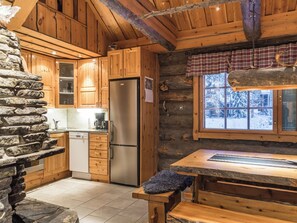 A log cabin interior with a stone wall, wooden cabinets, a stainless steel refrigerator, and a window with a view of snow-covered trees. - Operonmukka a 8 by Interhome (Kittilä)