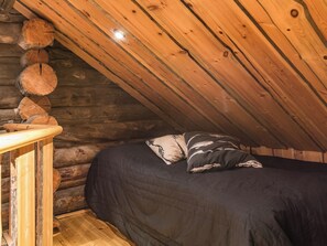 Quarto de cabana de madeira com teto de madeira, uma cama com cobertor escuro e mesa de cabeceira de madeira.