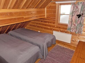 Quarto pequeno e aconchegante com paredes de madeira e teto inclinado, com duas camas cobertas com roupa de cama cinza e uma única janela com cortina.