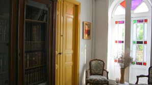 A room with a wooden bookshelf, a chair, a small table with a vase, and a stained glass window. - Mas de Bellegou B&B (Portiragnes)