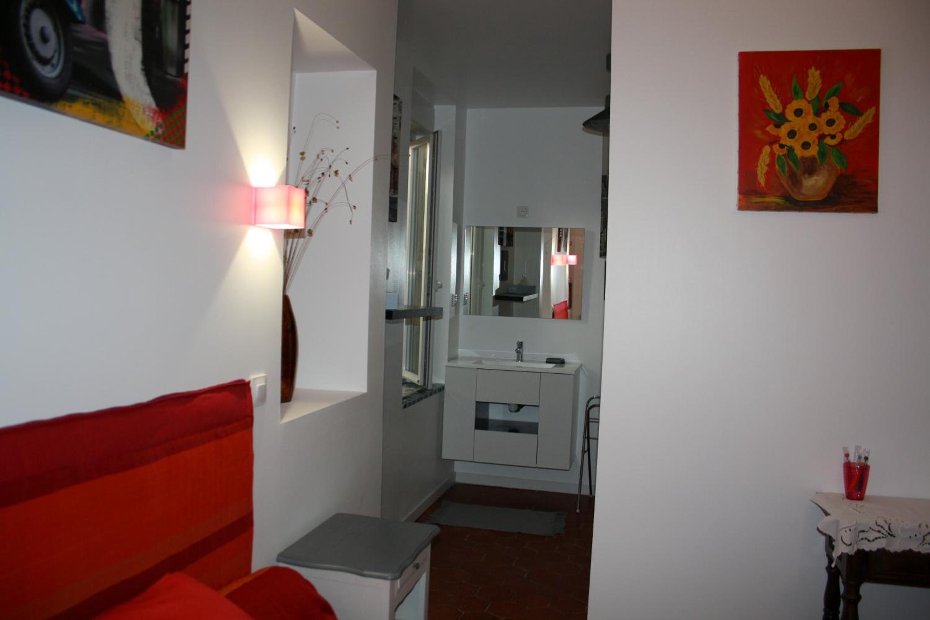 Una habitación de hotel con una cama roja, una mesita de noche pequeña, un tocador, un espejo, un lavamanos y un jarrón rojo sobre una mesa.