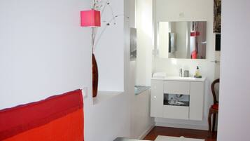 A bedroom with a red bed, a small bedside table, a mirror above the sink, and a chair.
