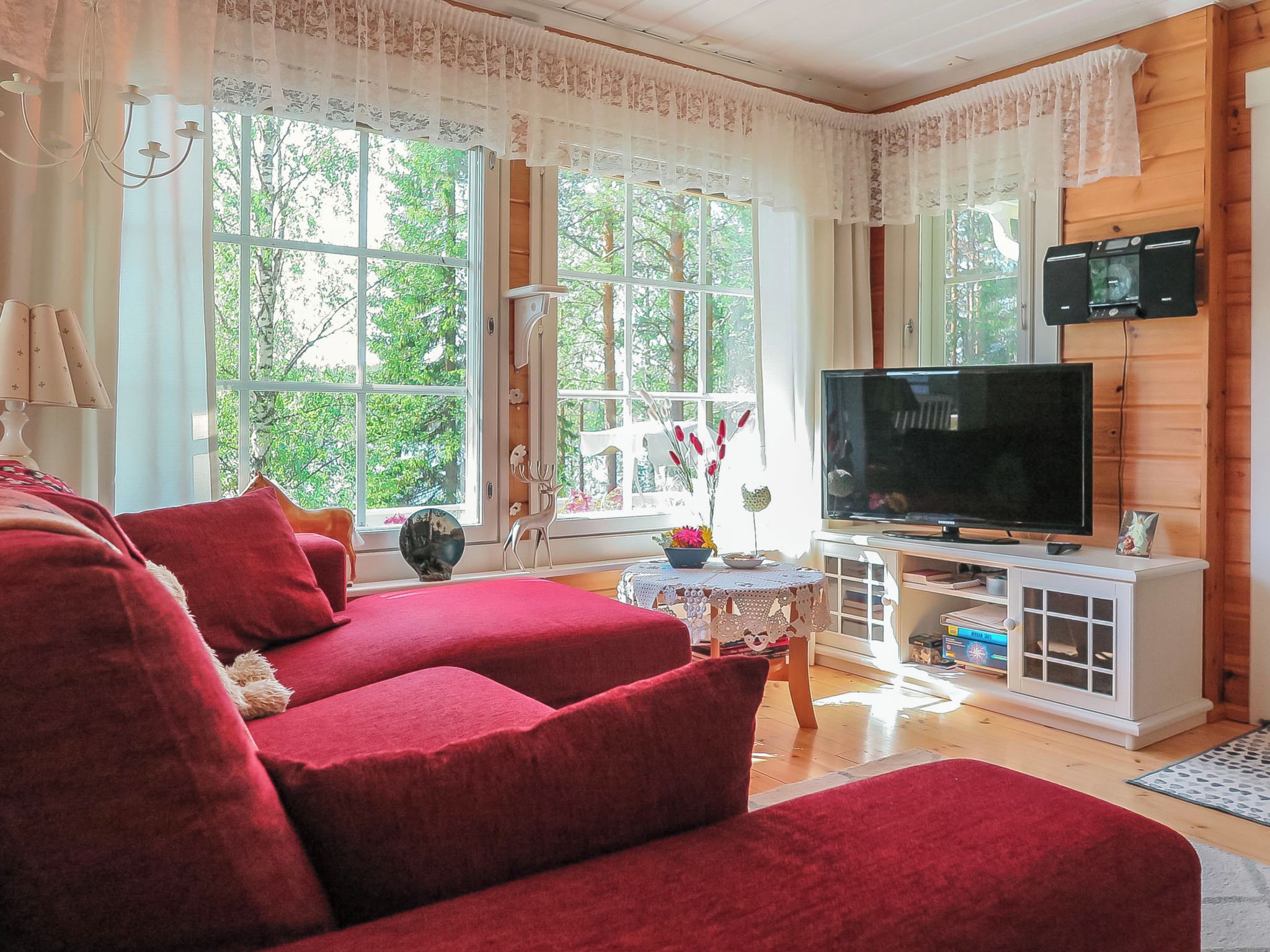 A living room with a red sofa, a television on a white cabinet, and large windows with lace curtains.