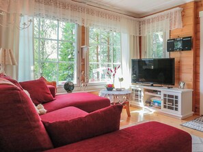 Ein Wohnzimmer mit einem roten Sofa, einem Fernseher auf einem weißen Schrank und großen Fenstern mit Spitzenvorhängen.