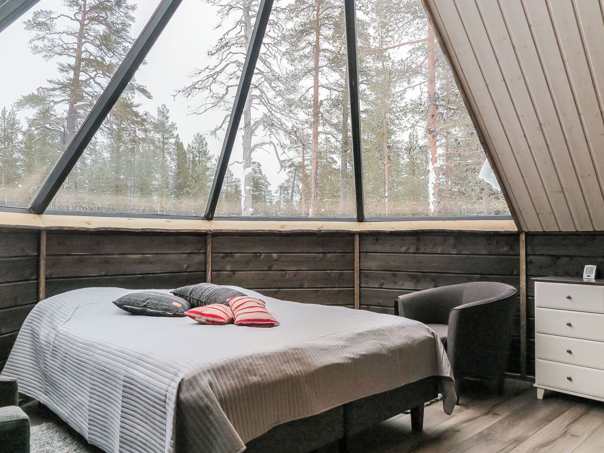 Un dormitorio con cama, almohadas, una silla, cómoda y ventana con vista a árboles.