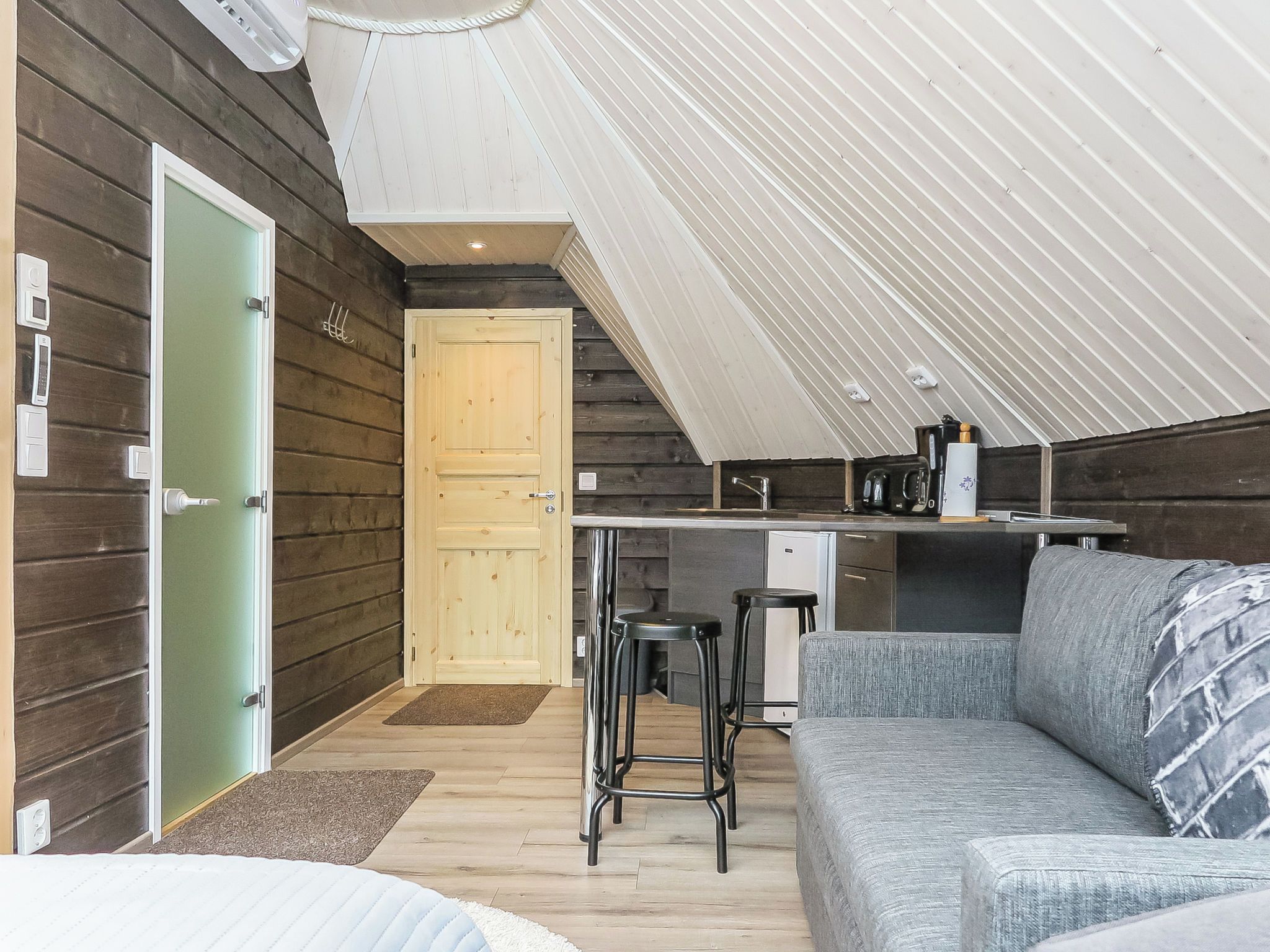 Un intérieur chaleureux avec une façade en bois, une kitchenette, un coin salon avec un canapé et des tabourets de bar, et une porte donnant sur une autre pièce.