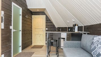 Un intérieur chaleureux avec une façade en bois, une kitchenette, un coin salon avec un canapé et des tabourets de bar, et une porte donnant sur une autre pièce.